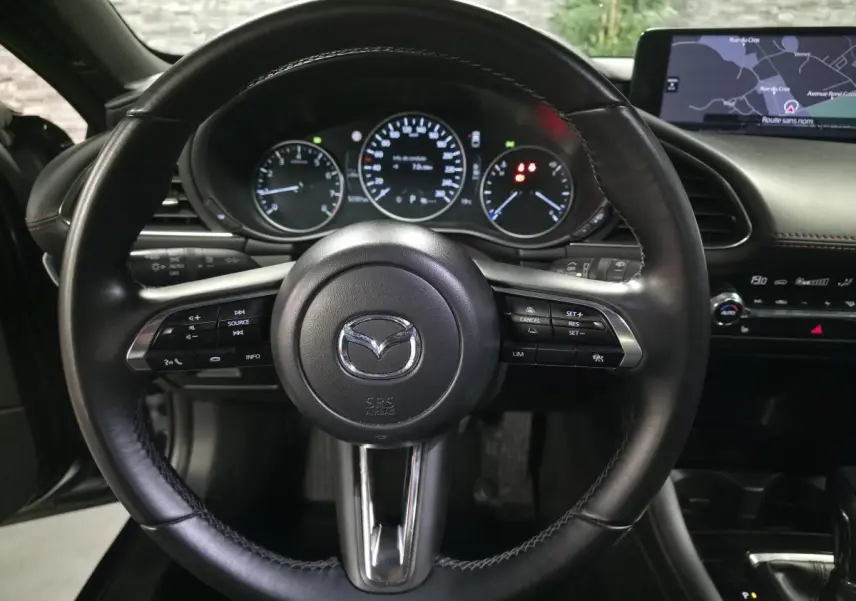 Vue intérieure centrée sur le volant cuir noir de la Mazda3 2024 avec tableau de bord et écran de navigation visibles.
