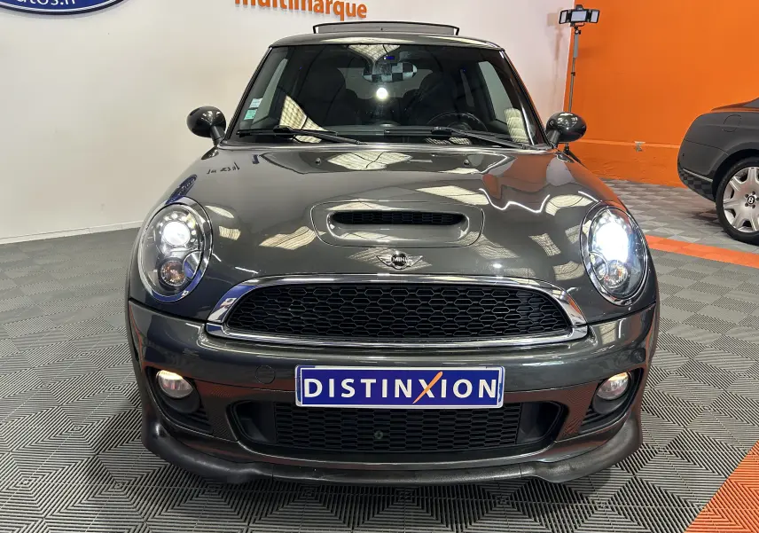 Vue avant d'une MINI Cooper gris foncé avec phares allumés, calandre noire et plaque Distinxion visible.