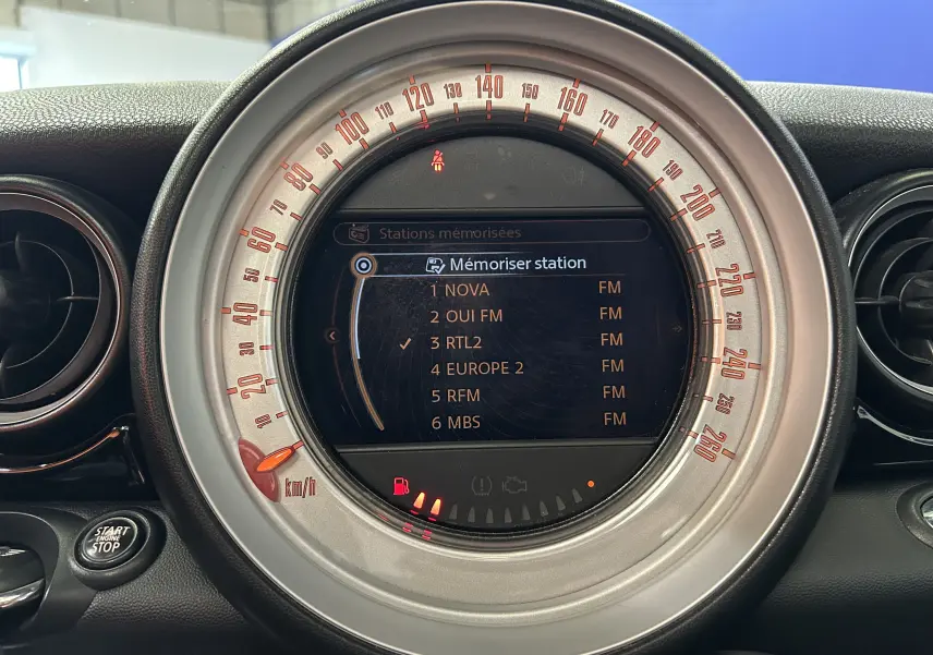 Gros plan sur le compteur central rond de la MINI Cooper gris, affichant l'écran radio avec stations FM mémorisées.