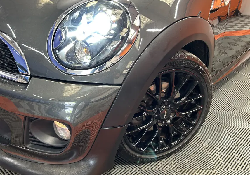 Gros plan sur l'avant droit gris métallisé d'une MINI Cooper avec phare allumé et jante noire brillante.