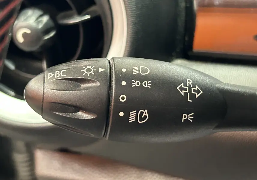 Gros plan sur la manette de clignotants noire du tableau de bord d’une MINI gris foncé, avec symboles blancs visibles.