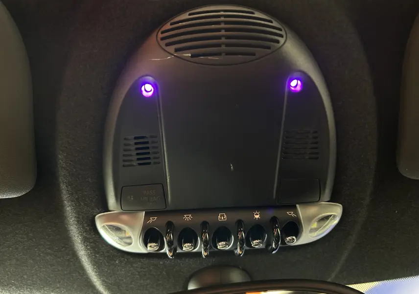 Vue en plongée sur le plafonnier noir de la MINI Cooper gris 2011 avec interrupteurs chromés et lumières violettes allumées.