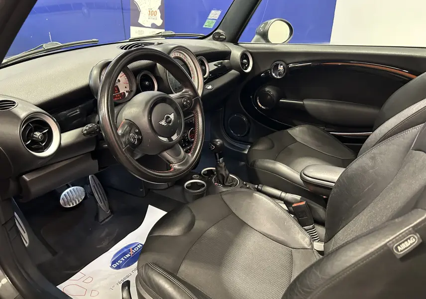 Vue intérieure côté conducteur de la MINI Cooper gris, avec volant cuir, sièges noirs et tableau de bord rond caractéristique.