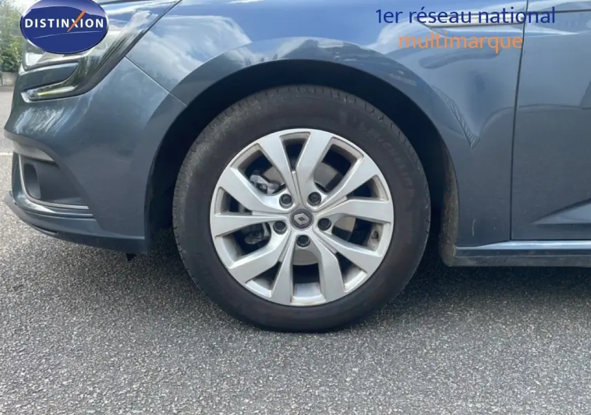 Gros plan sur la roue avant gauche d'une Renault Mégane gris titanium métal, avec jante alliage et partie avant du véhicule visible.