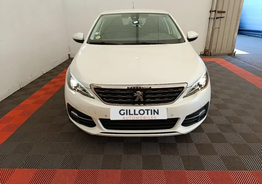 Peugeot 308 blanche vue de face dans un garage, avec calandre chromée et phares allumés.
