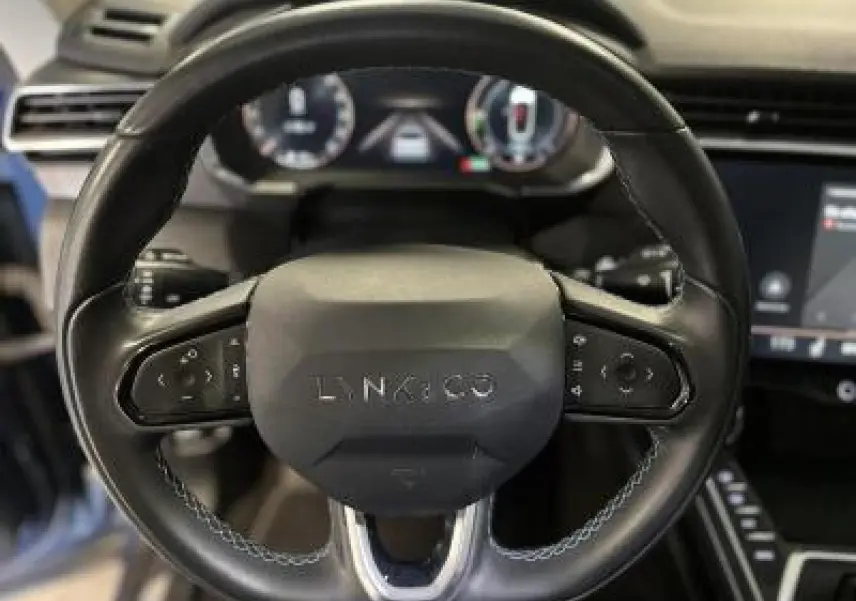 Vue rapprochée du volant noir du Lynk & Co 01 1.5 197 Hybride avec commandes intégrées et tableau de bord numérique flou.