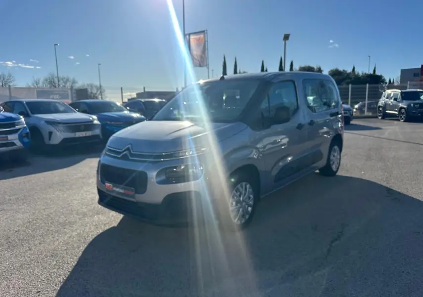 Citroën Berlingo gris clair vue 3/4 avant droit avec protections latérales noires, stationné en extérieur au soleil.