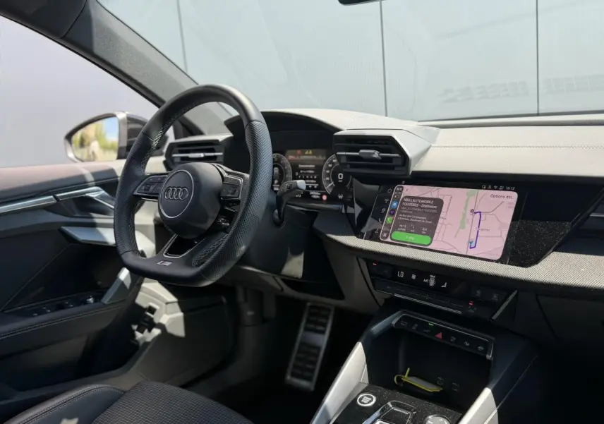 Vue intérieure côté conducteur de l'Audi A3 Sportback 2025, volant cuir noir et écran tactile de navigation allumé.