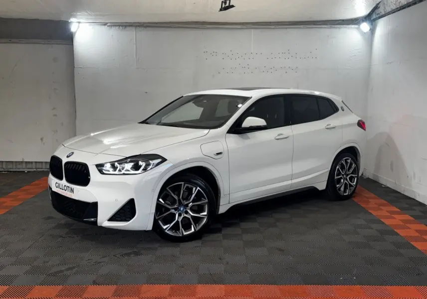 BMW X2 xDrive25e blanc en 3/4 avant droit, avec jantes alliage et toit ouvrant panoramique visible.