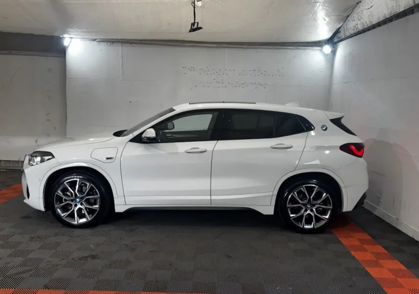 BMW X2 xDrive25e blanc en vue profil côté gauche dans un garage, avec jantes alliage et logo M Sport visible.