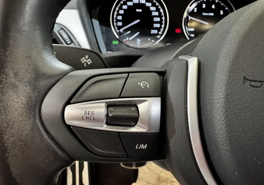 Gros plan sur les commandes de régulateur de vitesse à gauche du volant cuir noir d'une BMW X2 blanche, tableau de bord en arrière-plan.