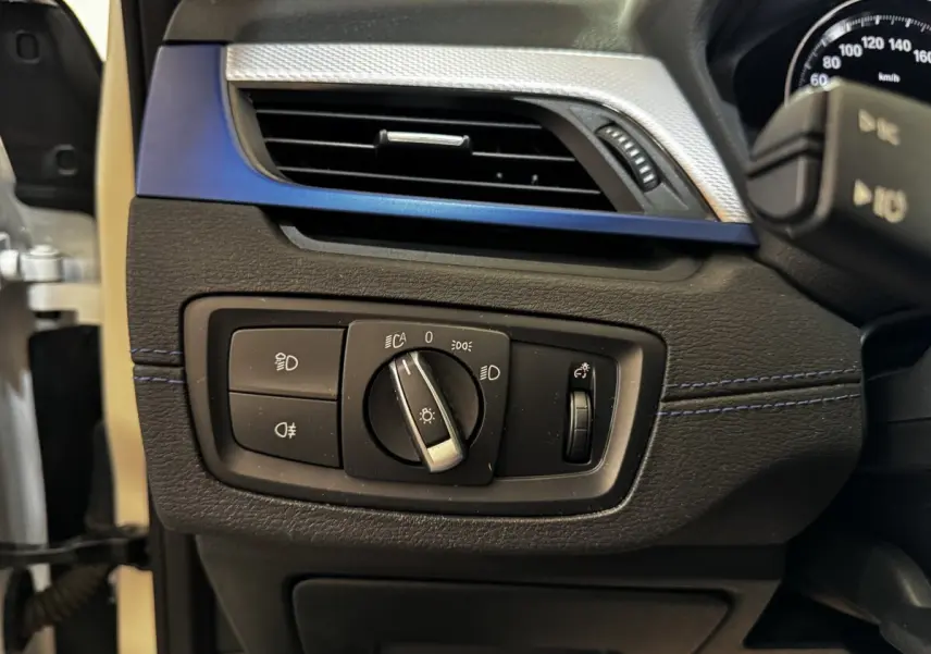 Gros plan sur le panneau de commande des phares à gauche du volant dans l’habitacle noir et bleu d’une BMW X2 blanche.