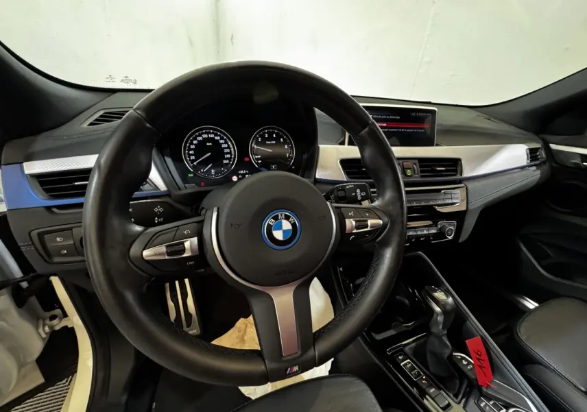 Vue intérieure centrée sur le volant cuir M et tableau de bord avec écran couleur de la BMW X2 blanc hybride.