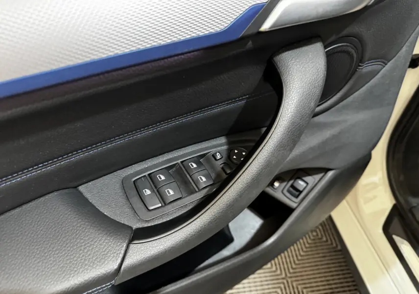 Gros plan sur la porte avant gauche noire avec commandes des vitres et poignée, BMW X2 blanc visible en arrière-plan.