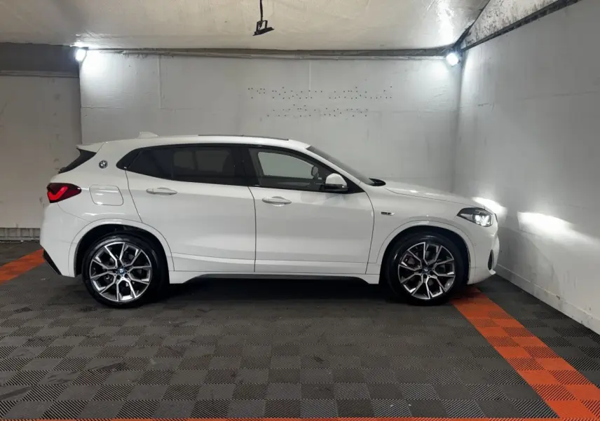 Vue de profil côté gauche d’un BMW X2 blanc avec jantes alliage et feux arrière LED allumés dans un parking fermé.