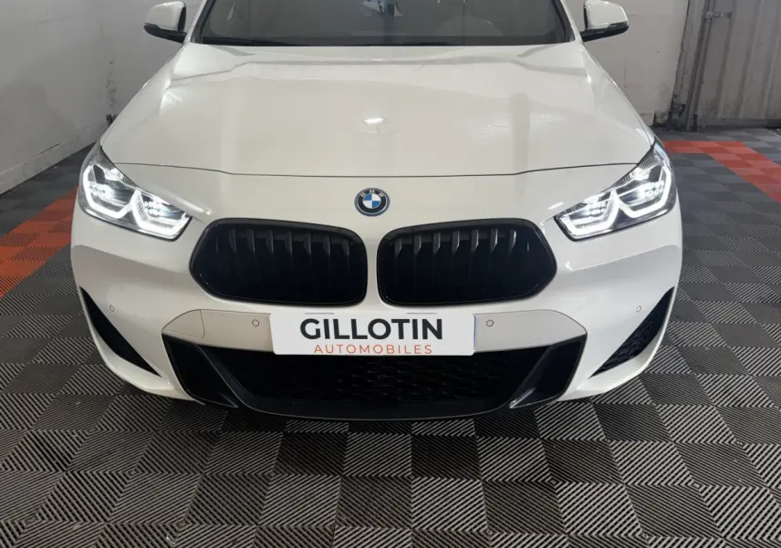 Vue avant d’une BMW X2 xDrive25e blanche avec calandre noire et phares LED allumés dans un garage.
