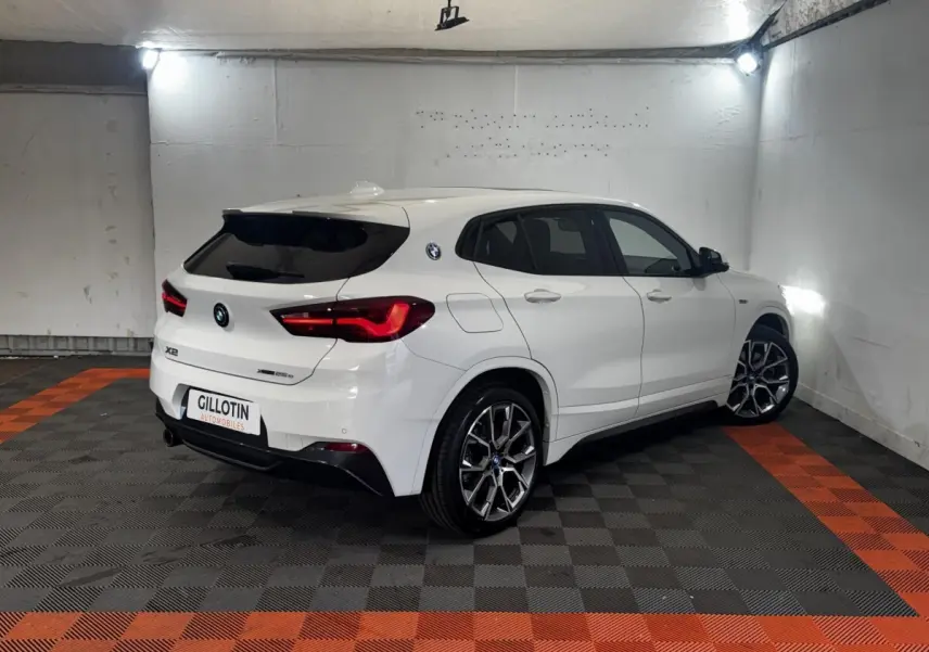 BMW X2 blanc vue 3/4 arrière droit dans un garage, avec jantes alliage et feux arrière LED allumés.