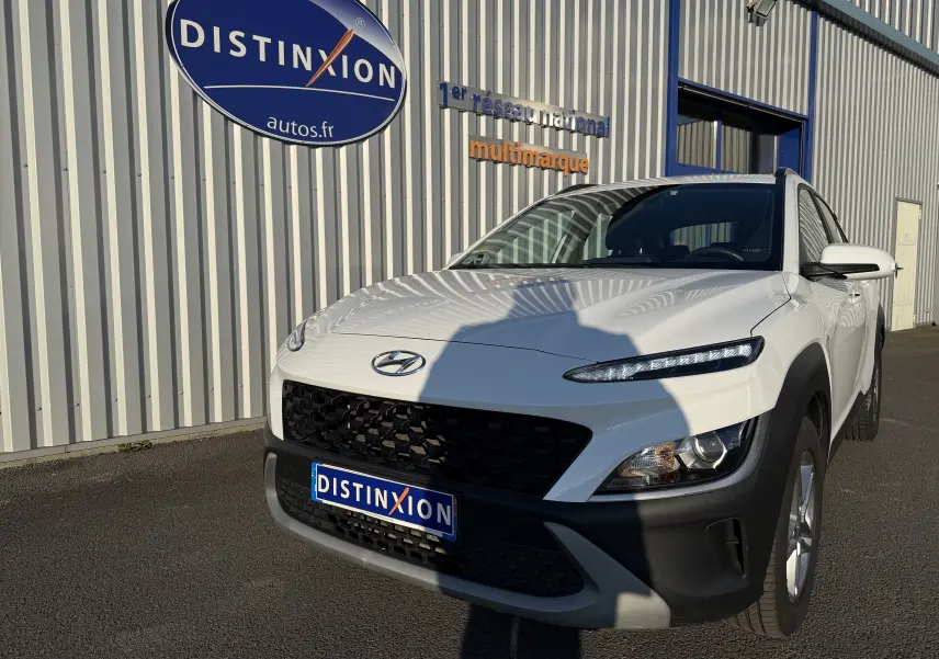 Hyundai Kona blanc Atlas en 3/4 avant droit, avec calandre noire et phares LED distinctifs, stationné devant un bâtiment.