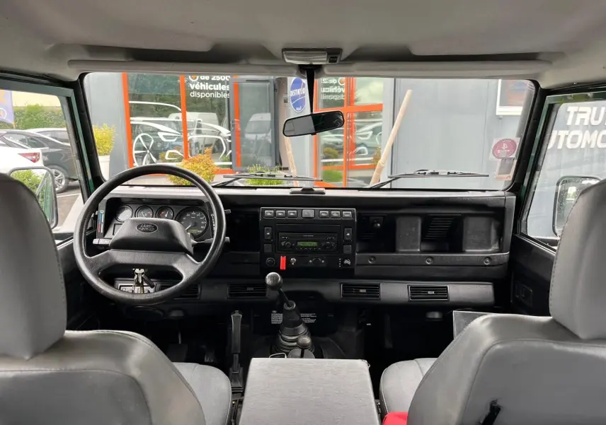 Vue intérieure avant du Land Rover Defender 90 Hard Top 2004, tableau de bord noir avec volant et levier de vitesses manuelle visibles.