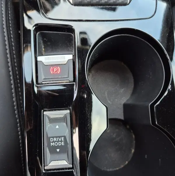 Gros plan sur la console centrale noire brillante du Peugeot 2008 Hybrid 145 e-DCS6 GT avec bouton frein à main et sélection drive mode.