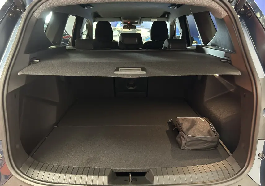 Coffre ouvert vu de l'arrière du Dacia Bigster bleu indigo, avec cache-bagages et sac noir posé à droite.