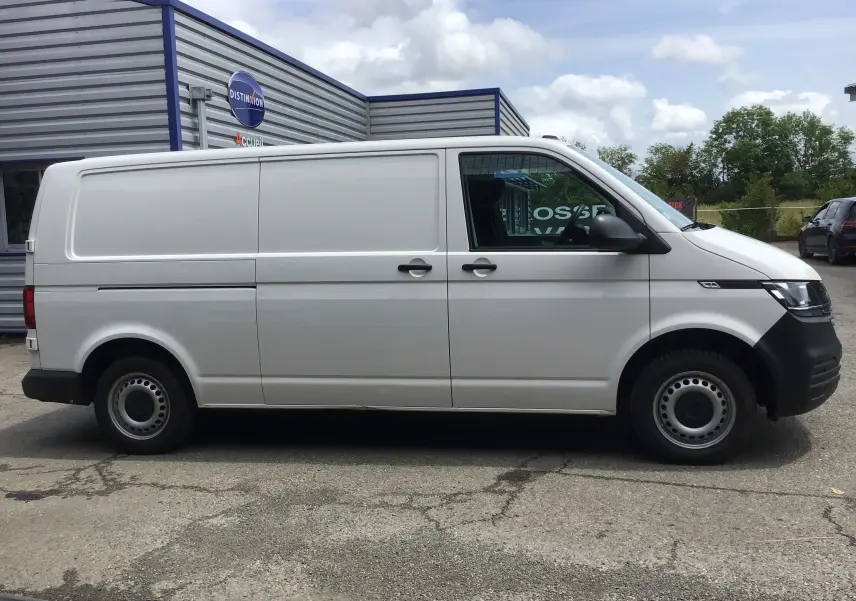 Profil côté gauche d’un utilitaire Volkswagen Transporter 6.1 blanc, version L2H1 avec portes latérales coulissantes, stationné en extérieur.