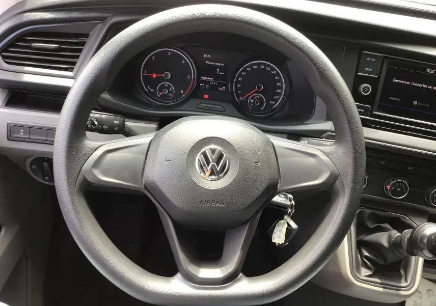 Vue rapprochée du volant noir et du tableau de bord du Volkswagen Transporter 6.1 blanc avec boîte manuelle et écran multimédia.