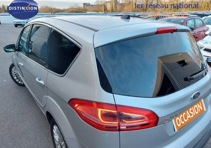 Vue 3/4 arrière droite d'un Ford S-MAX gris clair métal avec feux arrière allumés sur parking extérieur.