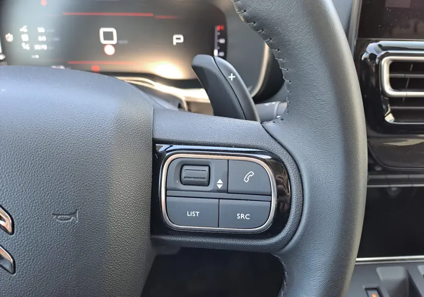 Gros plan sur les commandes gauche du volant cuir du Citroën C5 Aircross 2024 hybride, avec tableau de bord numérique en arrière-plan.