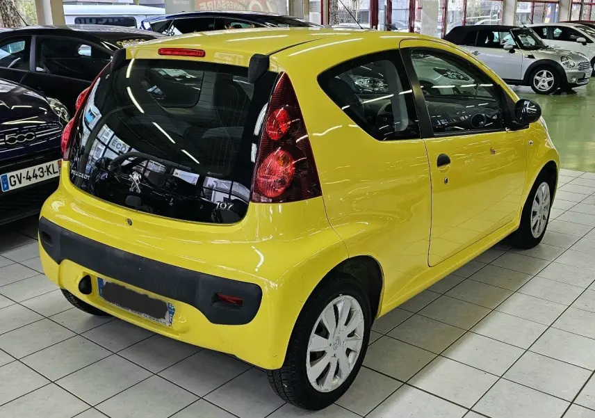 Vue 3/4 arrière droite d'une Peugeot 107 jaune vif en intérieur, avec feux arrière verticaux et pare-chocs noir.