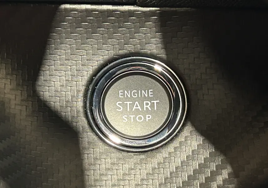 Gros plan sur le bouton moteur Start/Stop entouré d'un cerclage chromé sur un fond noir à motif carbone.