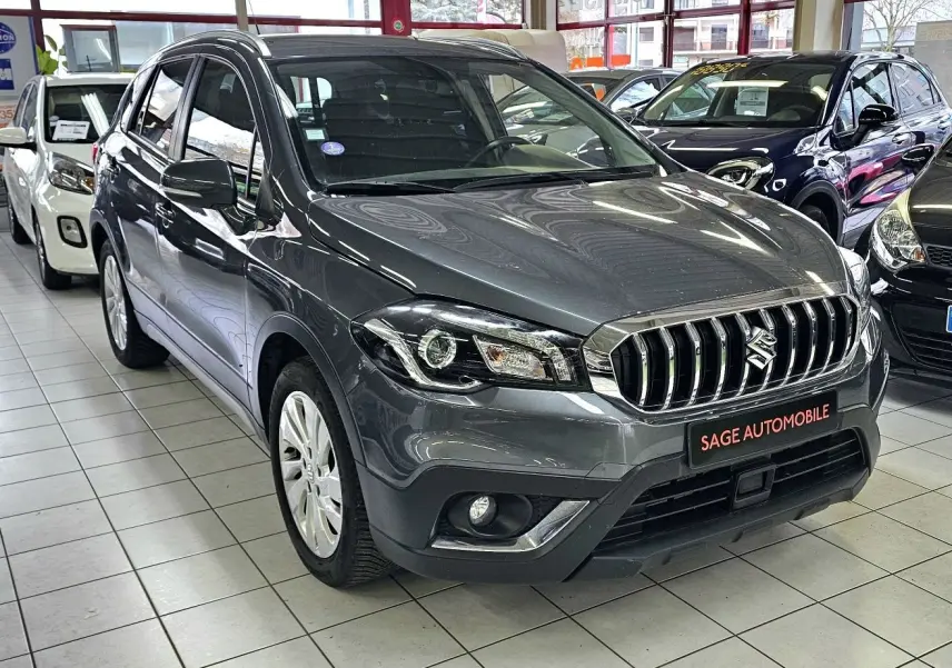 Suzuki SX4 S-Cross gris en 3/4 avant droit, avec calandre chromée et antibrouillards intégrés, en showroom.