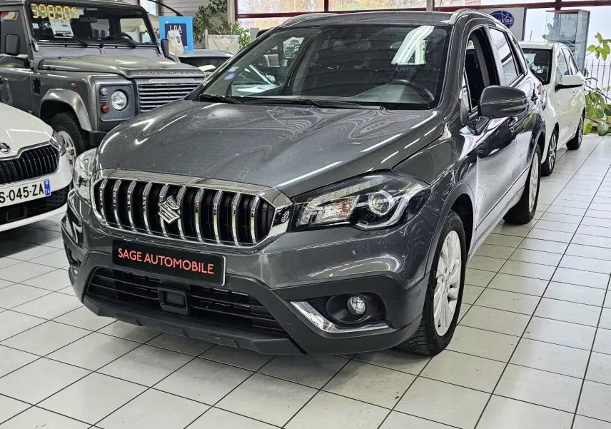 Suzuki SX4 S-Cross gris vu de face droite en showroom, avec calandre chromée et phares LED visibles.