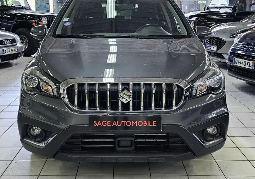 Vue frontale d'un Suzuki SX4 S-Cross gris exposé en showroom avec calandre chromée et phares allumés.
