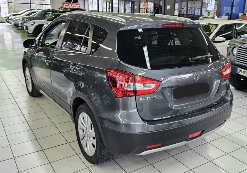 Suzuki SX4 S-Cross gris en 3/4 arrière droit, stationné en intérieur avec plusieurs autres véhicules visibles.