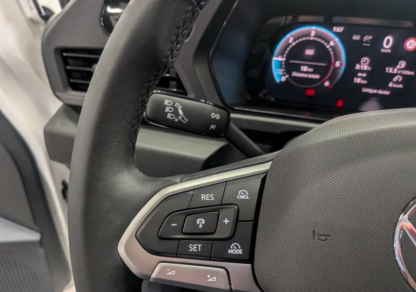 Gros plan sur le volant noir du Volkswagen Caddy blanc 2025, avec commandes de régulateur et tableau de bord numérique visible.