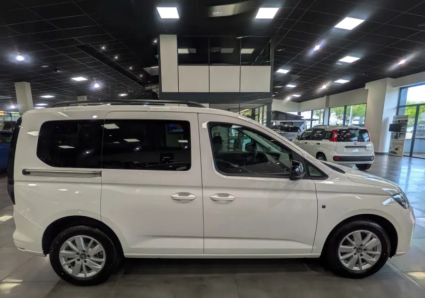 Vue de profil côté gauche d'un Volkswagen Caddy blanc 2025 en showroom, avec vitres arrière teintées et jantes alliage.