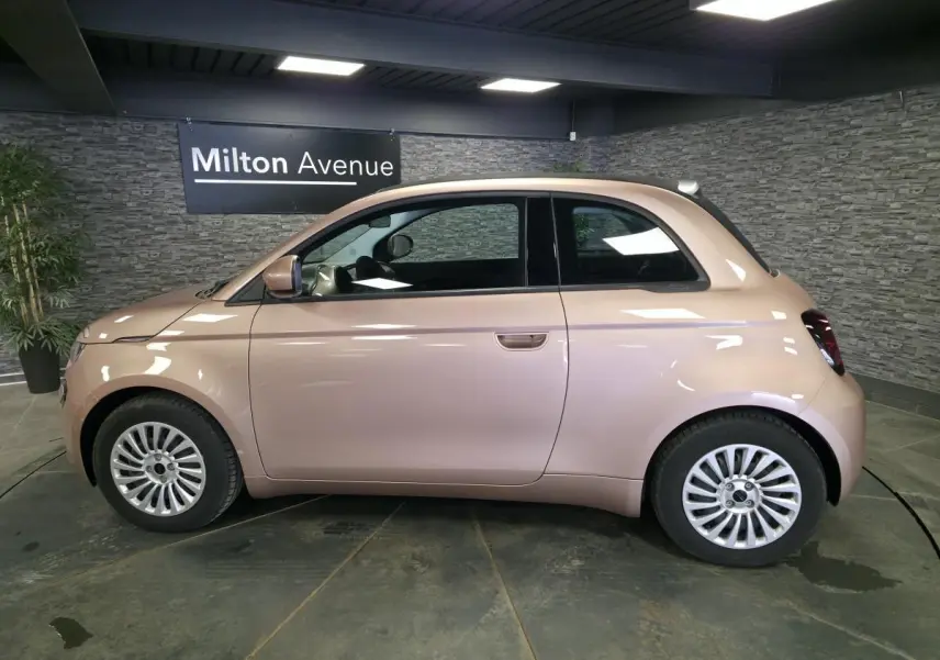 Vue de profil côté gauche d'une FIAT 500 Cabriolet électrique rose métallisée avec jantes acier 15 pouces en intérieur showroom.