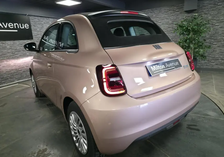 Vue 3/4 arrière droite d'une Fiat 500 Cabriolet électrique rose métallisé avec capote noire en intérieur showroom.