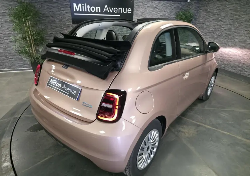 Vue 3/4 arrière droite d'une Fiat 500 Cabriolet électrique rose métallisé avec capote noire partiellement ouverte.