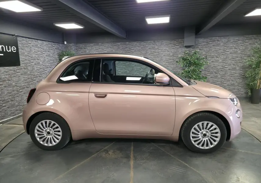 Vue de profil côté gauche d'une FIAT 500 Cabriolet électrique 2023 rose métallisée avec jantes acier et capote noire fermée.