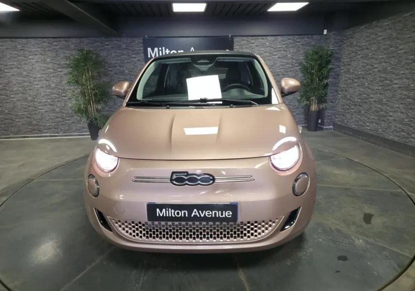 Vue avant d'une FIAT 500 Cabriolet électrique rose métallisée avec phares allumés en intérieur showroom.