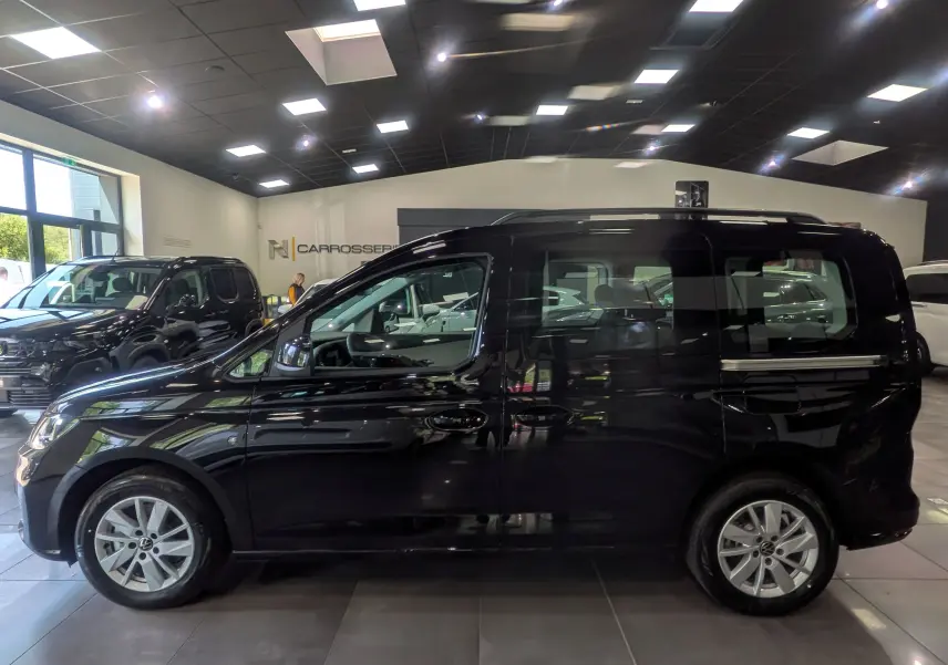 Vue de profil côté gauche d'un Volkswagen Caddy noir 2025 en showroom, avec jantes alliage et vitres teintées.