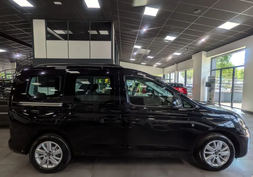 Vue de profil côté gauche d'un Volkswagen Caddy noir 2025 en showroom, avec jantes alliage visibles.