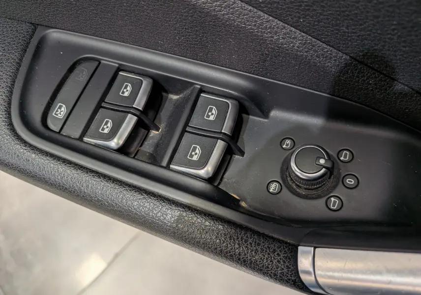 Gros plan sur les commandes de lève-vitres et réglage des rétroviseurs côté conducteur en plastique noir d'un Audi Q2 gris.