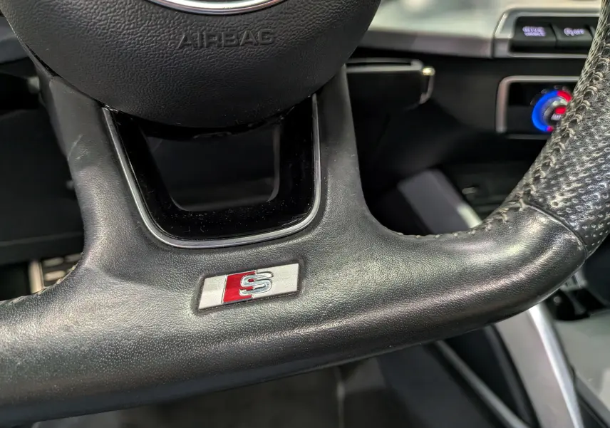 Gros plan sur le volant en cuir noir avec logo S de l'Audi Q2 1.4 TFSI S-Line, détails du tableau de bord en arrière-plan.