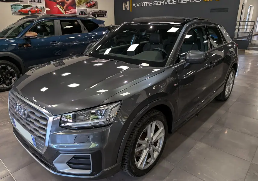 Audi Q2 gris métallisé vue 3/4 avant droit en intérieur, avec jantes alliage et toit ouvrant panoramique.
