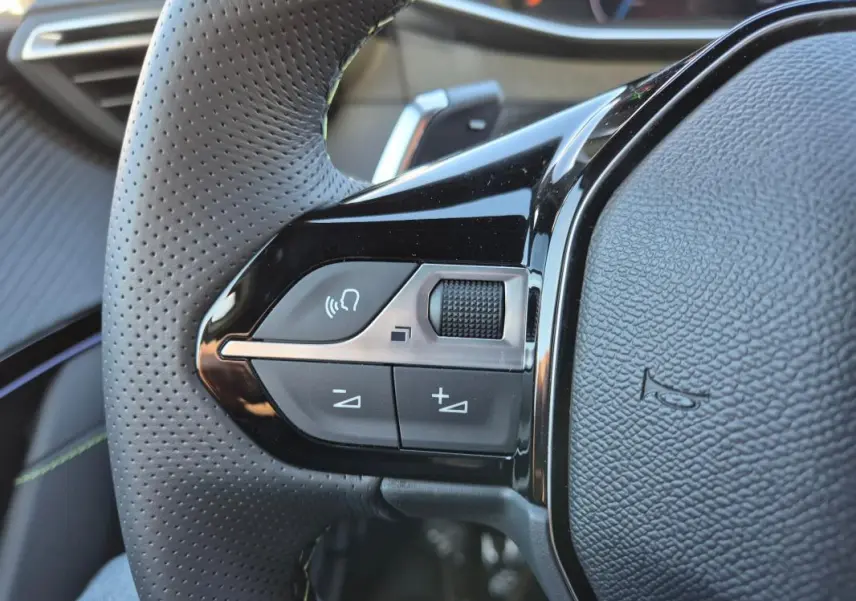 Gros plan sur le côté gauche du volant perforé noir de la Peugeot 208 Hybrid 136 e-DCS6 GT avec commandes audio intégrées.