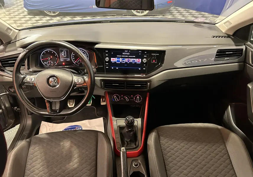 Vue intérieure avant de la Volkswagen Polo grise 2018, avec tableau de bord moderne et console centrale rouge autour du levier de vitesses manuel.