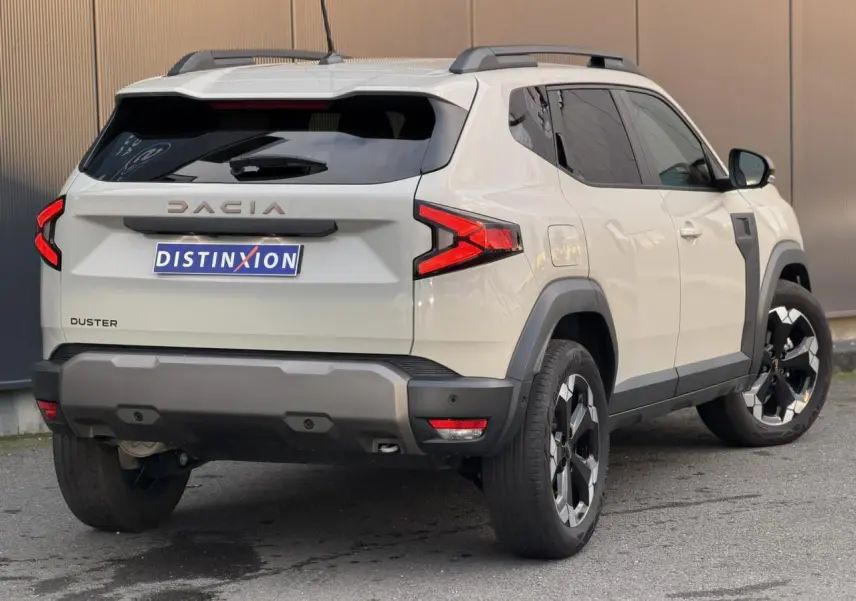 Vue 3/4 arrière droite du Dacia Duster beige Sandstone avec jantes alliage et barres de toit noires.